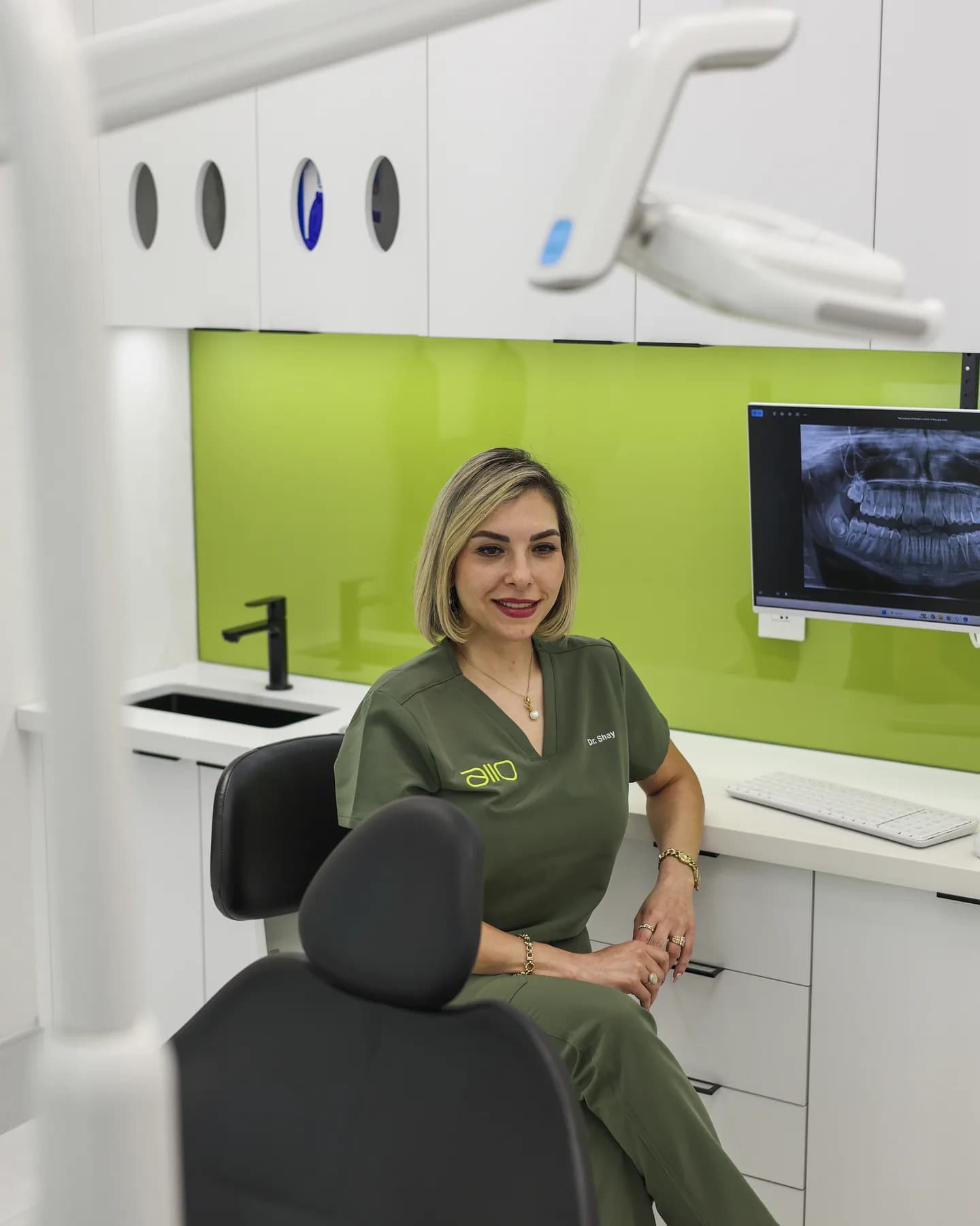 Dr. Shay Rahmatian seated beside the dental chair in a treatment room, with a panoramic X-ray on the screen behind her