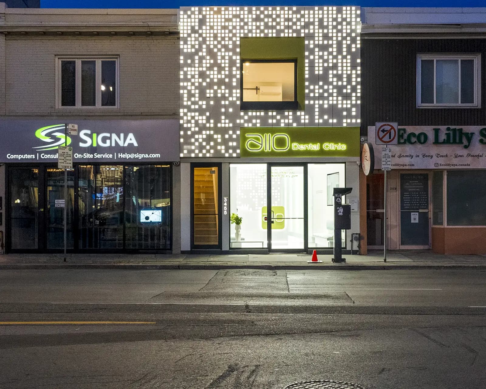 The illuminated perforated facade of Allo Dental Clinic at dusk, between its neighbouring storefronts on Yonge Street