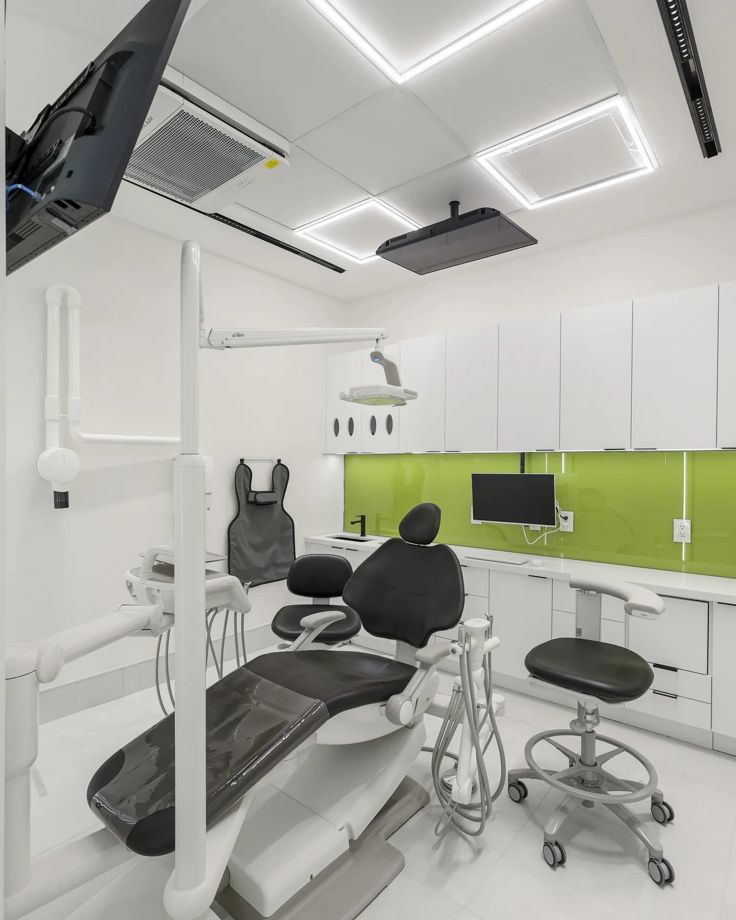 A treatment room at Allo Dental Clinic with a dark dental chair, chairside monitor, and olive-green glass backsplash
